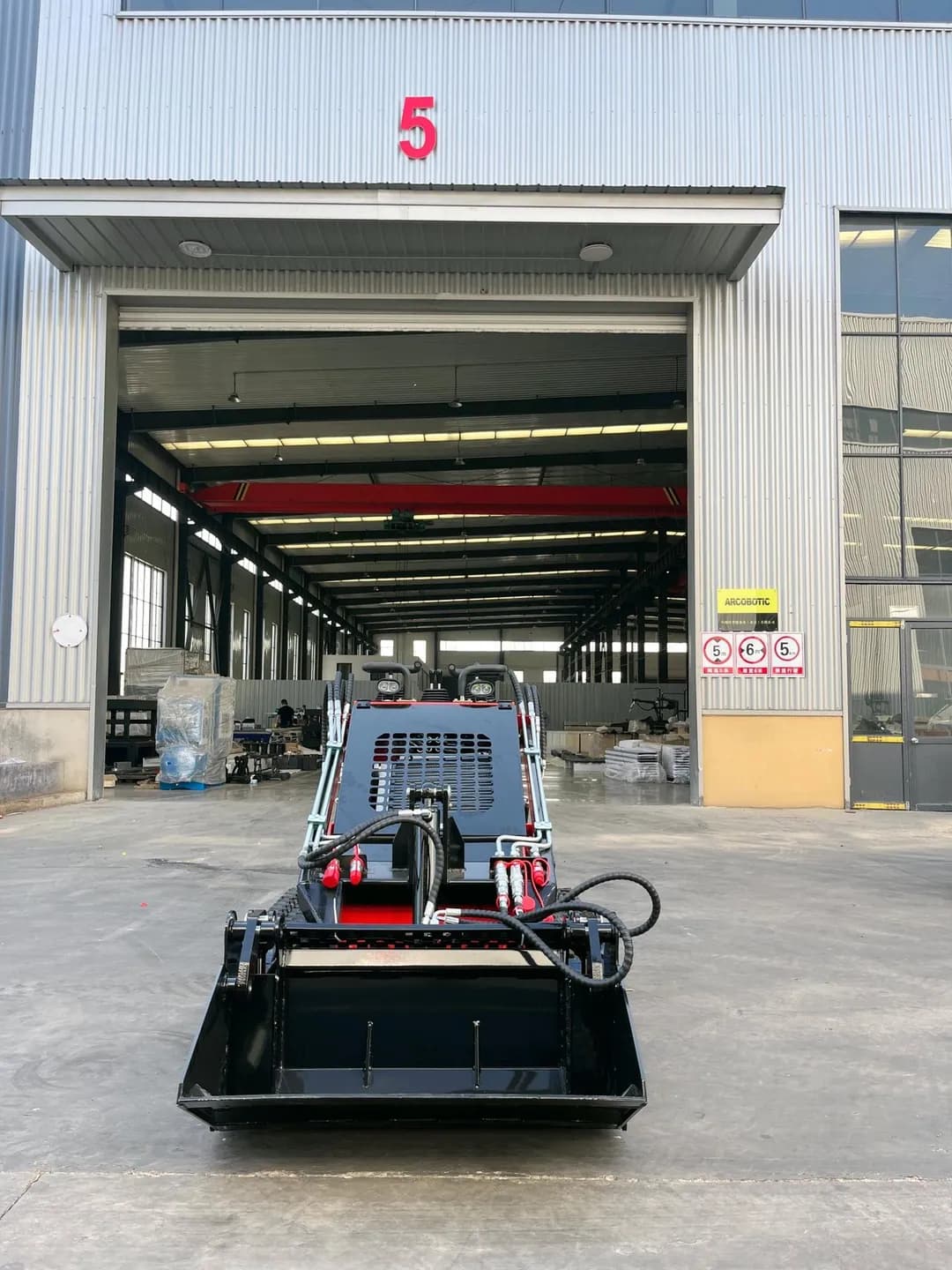 Front of a red stand-on loader with bucket lowered outside a workshop